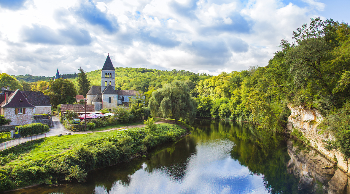 A river running past lush greenery and a small picturesque town with a turreted building